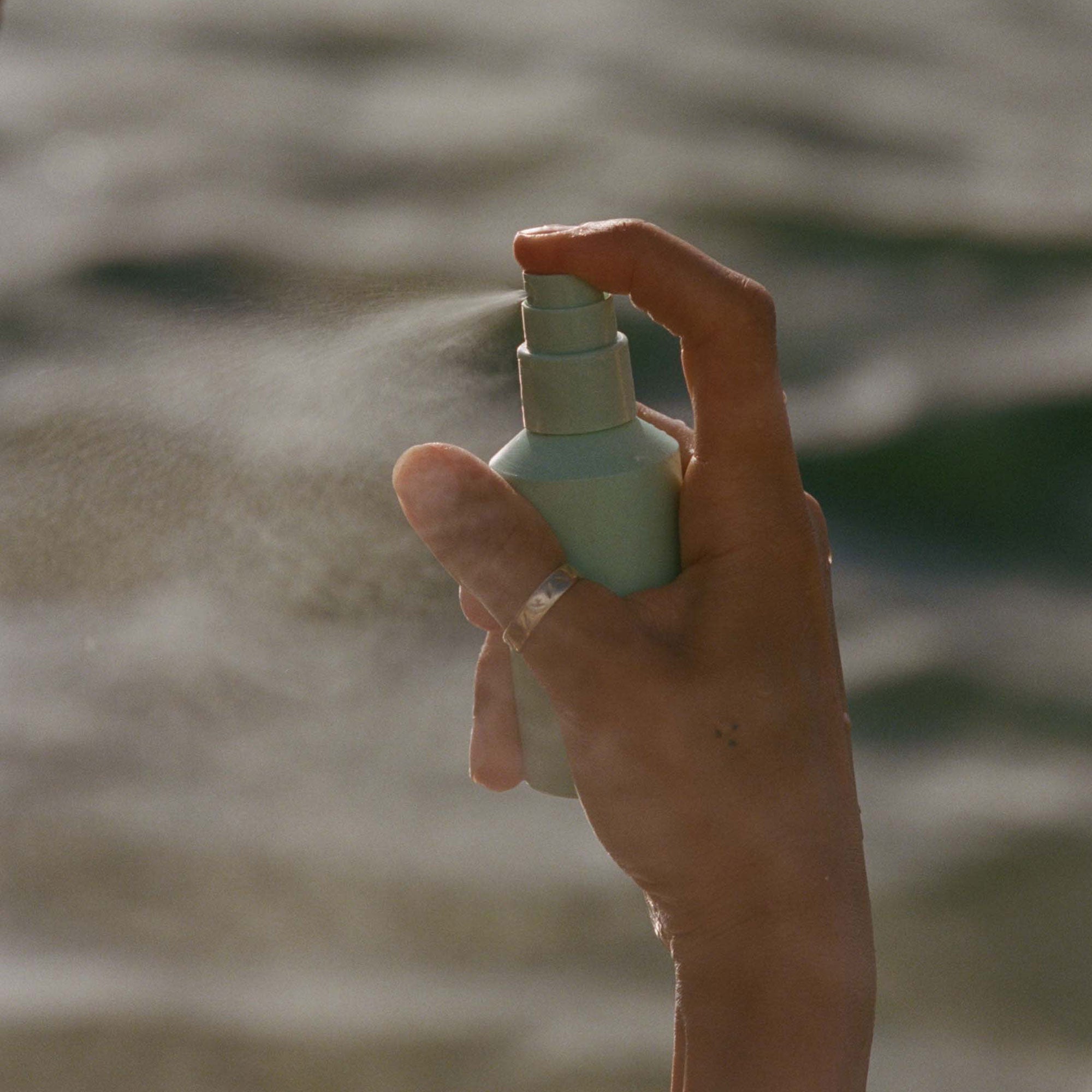 Hand spraying mist from a small bottle against a blurred water background